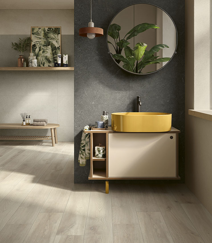 Modernes Badezimmer mit Oberflächen aus den Kollektionen Surround Outline Soft, Draft und Borealis Inari, mit dunkler Wand, gelbem ovalem Waschbecken, schwebendem Waschtisch, großem Rundspiegel, dekorativen Pflanzen und hellem Boden in Holzoptik.