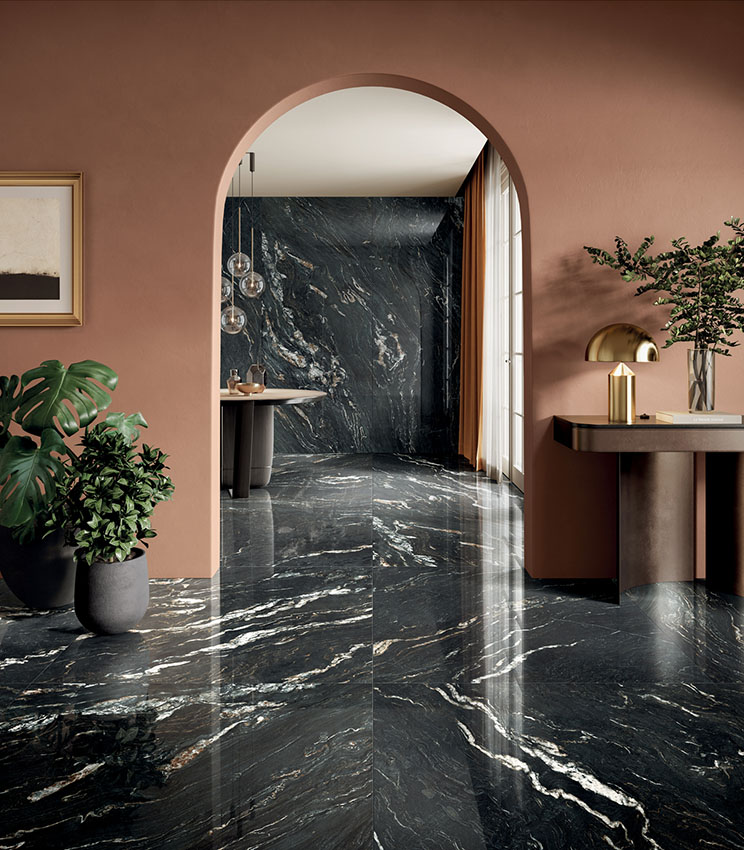 Modernes Interieur mit Oberflächen aus der Kollektion Perpetual Belvedere Dark Lux, geprägt von großformatigen dunklen Platten in Marmoroptik mit heller Äderung und glänzender Oberfläche. Ergänzt wird der Raum durch terrakottafarbene Wände, reduzierte Möbel, dekorative Pflanzen, goldfarbene Leuchten und einen Rundbogen.