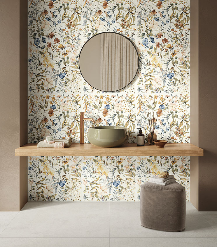 Modernes Badezimmer mit Oberflächen aus den Kollektionen La Matière Talc und La Fleur, mit floraler Wandgestaltung in natürlichen Farbtönen, olivgrünem Waschbecken, Holzablage, rundem Spiegel und gepolstertem Hocker auf hellem Boden.