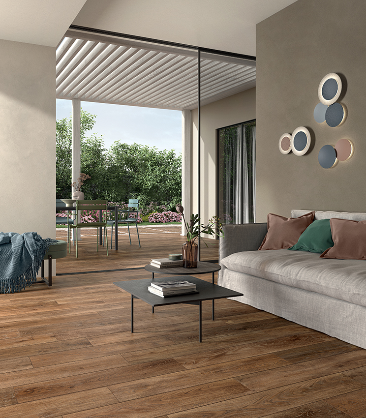 Modernes Wohnzimmer mit Bodenbelag aus der Kollektion Kairos Zoi, geprägt von warmen Oberflächen in Holzoptik, einem großen hellen Sofa, niedrigen Couchtischen und direktem Zugang zu einer möblierten Terrasse mit Blick ins Grüne.