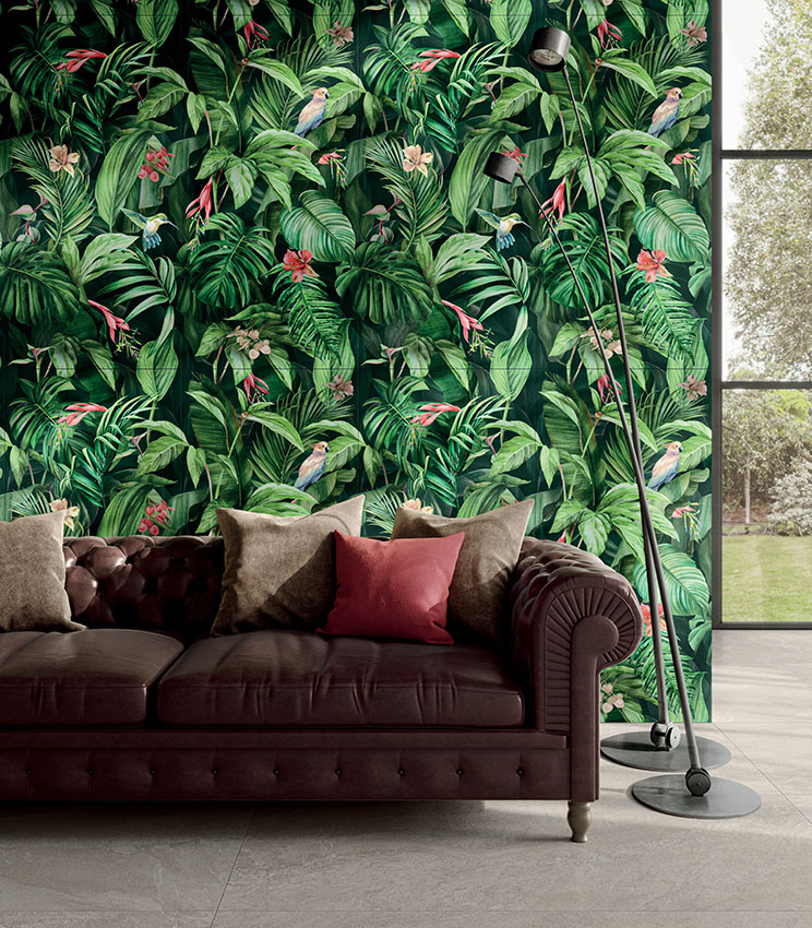 Modernes Wohnzimmer mit dekorativer Wandverkleidung aus der Kollektion Glam Vision, mit tropischem Muster aus Blättern, Blumen und Vögeln, dunklem Ledersofa mit Kissen in neutralen und bordeauxroten Tönen, schwarzen Stehleuchten und großer Fensterfront mit Blick in den Garten.