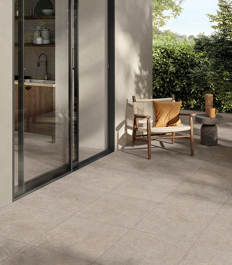 Moderne Terrasse mit Bodenbelag aus der Kollektion Surround Row, geprägt von großformatigen Platten in beigefarbener Steinoptik, einem Sessel aus Holz und Geflecht, einem skulpturalen Beistelltisch und üppiger Vegetation im Hintergrund. Große Glasflächen verbinden den Außenbereich mit der Küche im Inneren.