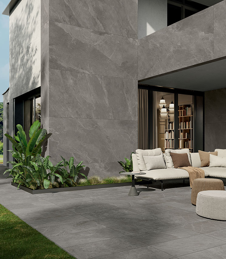 Außenansicht einer Villa mit modernem, streng geometrischem Design, teilweise mit großformatigen Keramikplatten in Steinoptik verkleidet, mit möbliertem Patio und einem Boden aus 20 mm starkem Feinsteinzeug in Steinoptik.