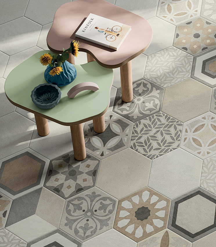 Detailansicht eines Bodenbelags mit Oberflächen aus den Kollektionen La Matière Hexa Talc Sable und Hexa Chic, mit sechseckigen Fliesen in neutralen Farbtönen, kombiniert mit geometrischen und floralen Dekormustern. Zwei weich geformte Tische in Grün und Rosa ergänzen die Szene mit Vasen und kleinen Dekorationsobjekten.