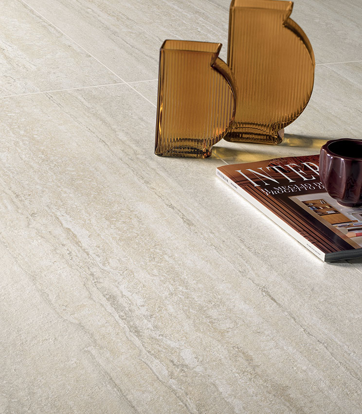 Detailansicht eines Bodenbelags aus der Kollektion Perpetual Travertino Ivory, geprägt von Oberflächen in Travertinoptik in hellen Beige- und Elfenbeintönen. Ergänzt wird die Szene durch zwei bernsteinfarbene Glasvasen, eine Zeitschrift und eine dunkle Tasse.