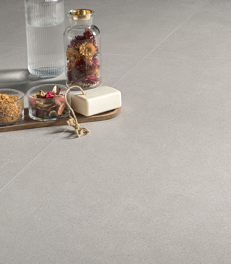 Detailansicht eines Bodenbelags aus der Kollektion Formula Form Ash, geprägt von einer hellen, gleichmäßigen Oberfläche mit materieller Optik. Ergänzt wird die Szene durch eine Komposition mit Glasflasche, Vase mit Trockenblumen, kleinen Behältern und einem Stück Seife auf einem Holztablett.