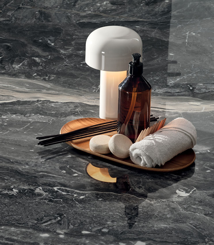 Detailansicht einer Oberfläche aus der Kollektion Perpetual Orobico Blend Lux, geprägt von dunkler Marmoroptik mit heller Äderung und glänzender Oberfläche. Ergänzt wird die Szene durch ein Holztablett mit Flakon, zusammengerolltem Handtuch, kleinen Accessoires und einer weißen Tischleuchte.