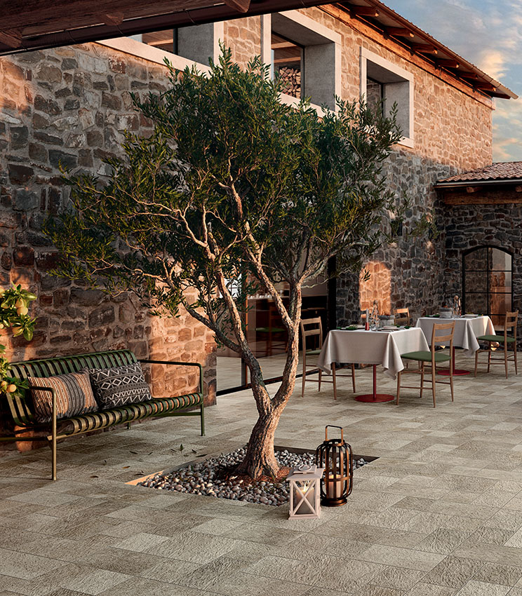 Restaurantterrasse mit Bodenbelag aus der Kollektion Pierre des Rêves Aurore, geprägt von Oberflächen in Steinoptik in natürlichen Farbtönen. Der von Steinmauern umgebene Bereich ist mit Holztischen und -stühlen, einer Bank, dekorativen Laternen und einem Olivenbaum in der Mitte ausgestattet.
