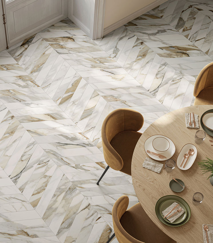 Moderner Essbereich mit Bodenbelag aus der Kollektion Perpetual Venato Gold Soft Chevron, geprägt von einem Chevron-Muster in weißer Marmoroptik mit grauer und goldener Äderung. Der Raum ist mit einem runden Tisch aus hellem Holz, ockerfarbenen Polsterstühlen und einer Tischdekoration in neutralen Farbtönen eingerichtet.