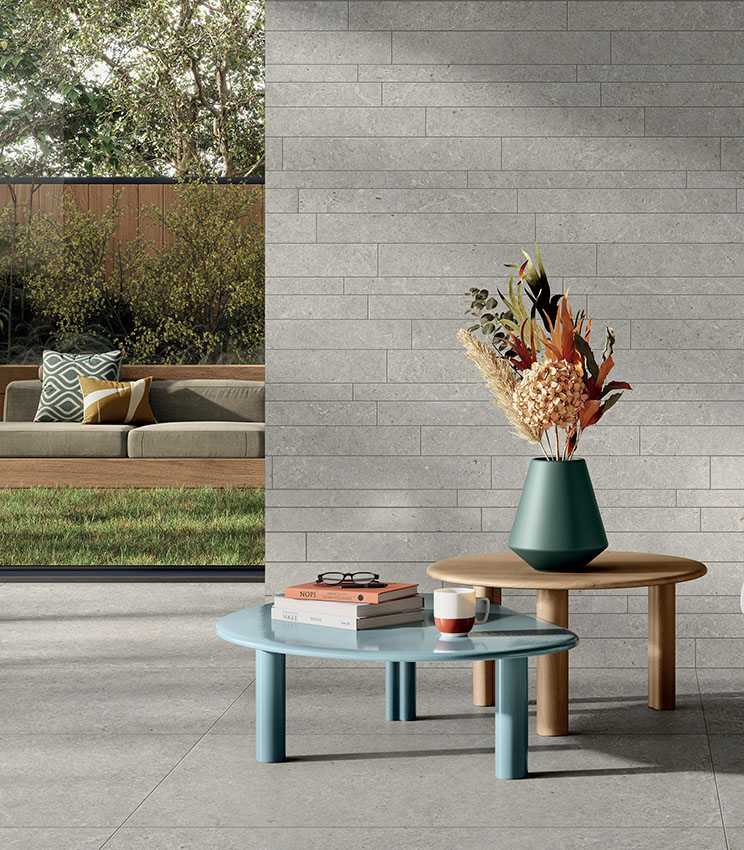 Modernes Wohnzimmer mit Oberflächen aus den Kollektionen Surround Shape und Shape Muretto Soft, mit grauem Boden in Steinoptik und einer Wandverkleidung aus abgestimmten rechteckigen Elementen. Der Raum ist mit schlichten Couchtischen, einer Vase mit Blumenarrangement und einer großen Öffnung zum Garten mit Außensofa eingerichtet.