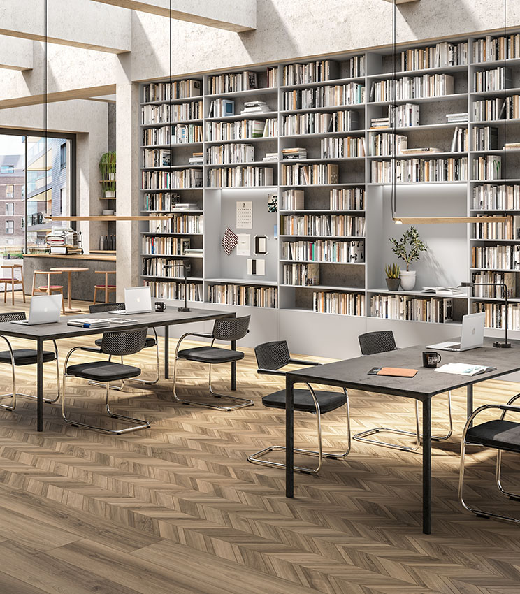 Moderne Bibliothek und Arbeitsbereich mit Bodenbelag aus den Kollektionen Nuance Cendre und Nuance Cendre Chevron, geprägt von Oberflächen in Holzoptik in natürlichen Farbtönen und einer geometrischen Verlegung. Der Raum ist mit großen Bücherregalen, Arbeitstischen mit schwarzen Stühlen, Laptops, dekorativen Pflanzen und großen Fenstern ausgestattet, die viel Tageslicht hereinlassen.