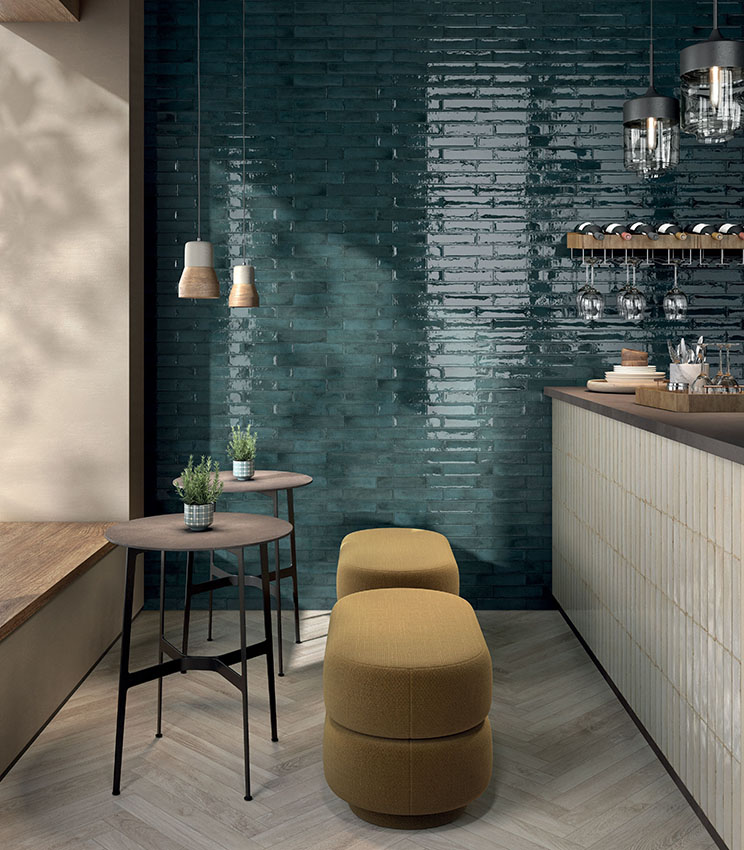 Moderner Barbereich mit Oberflächen aus den Kollektionen Workshop Berry, Workshop Opal und Kairos Imera, geprägt von einer glänzenden petrolgrünen Wand, einer hell verkleideten Theke, einem Boden in Holzoptik sowie kompakten Möbeln mit gepolsterten Hockern und hohen Tischen.