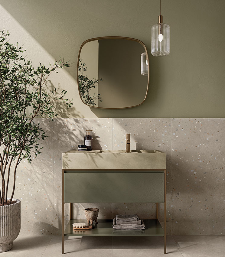 Modernes Badezimmer mit Oberflächen aus den Kollektionen Formula Form Rope und Seed Rope, geprägt von einer hellen Wandverkleidung mit kleinen farbigen Einschlüssen, salbeigrünen Wänden, einem passenden Waschtisch, einem großen Formspiegel, einer Pendelleuchte und einer dekorativen Pflanze.