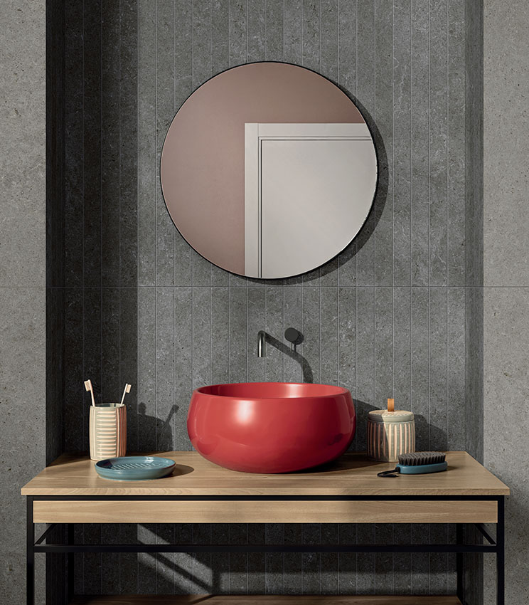 Modernes Badezimmer mit Oberflächen aus den Kollektionen Surround Shape Soft und Outline Carving, geprägt von grauen Verkleidungen in Steinoptik und einem zentralen Bereich mit vertikalem Linienmuster. Der Raum ist mit einem glänzend roten Waschbecken auf einer Holzplatte, einem runden Spiegel, dunkler Armatur und kleinen Badaccessoires ausgestattet.