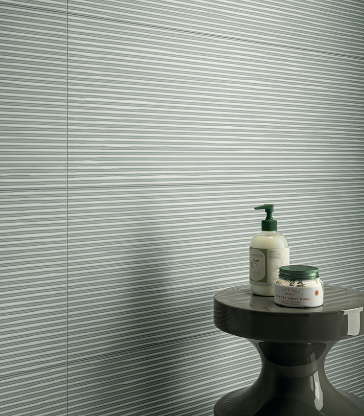 Detailansicht eines modernen Badezimmers mit Wandverkleidung aus der Kollektion Lullaby Linear Green, geprägt von einer hellgrünen Oberfläche mit horizontalem Linienmuster. Ergänzt wird die Szene durch einen dunklen skulpturalen Beistelltisch mit Flakons und kleinen Badprodukten.