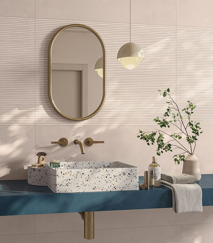 Modernes Badezimmer mit Oberflächen aus den Kollektionen Lullaby Fairytale Blush und Linear Blush, geprägt von puderrosa Wandverkleidungen mit Bereichen in linearer Reliefstruktur. Der Raum verfügt über ein rechteckiges Waschbecken in Terrazzo-Optik, eine blaue Ablage, einen ovalen Spiegel mit goldfarbenem Rahmen, passende Armaturen, eine Pendelleuchte und dekorative grüne Zweige.