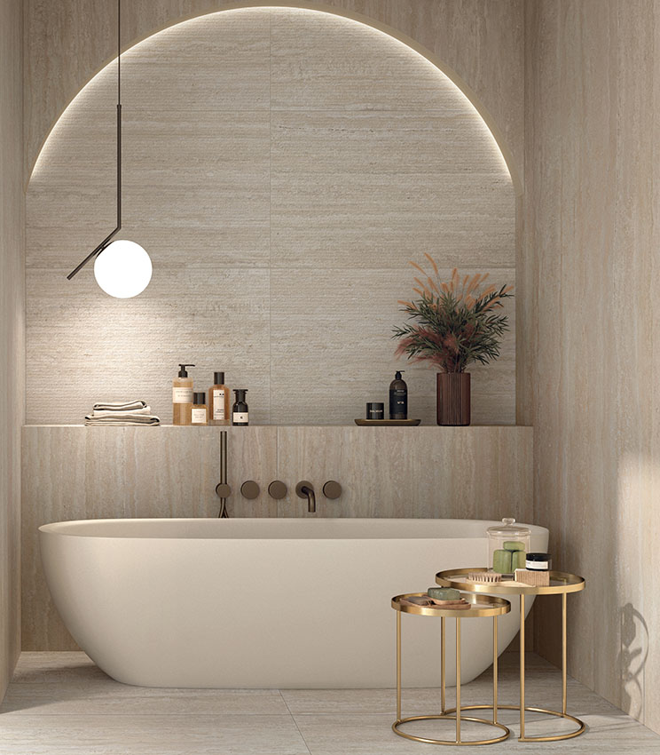 Modernes Badezimmer mit Oberflächen aus den Kollektionen Perpetual Travertino Ivory Edge, Travertino Ivory und Travertino Cream, geprägt von Travertinoptik in warmen, natürlichen Farbtönen. Der Raum verfügt über eine weiße freistehende Badewanne in einer hinterleuchteten Rundbogennische, integrierte Ablagen, eine Pendelleuchte und goldfarbene Metalltische mit Badaccessoires.