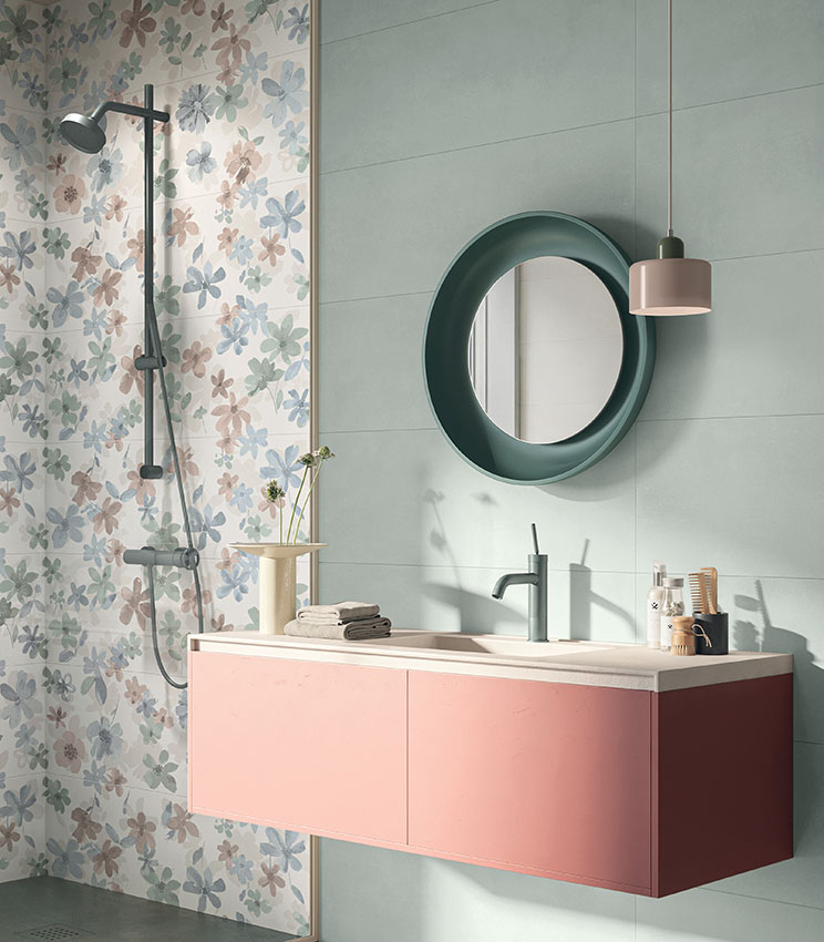Modernes Badezimmer mit Oberflächen aus den Kollektionen Lullaby Silent Green, Florè und Borealis Abisko, geprägt von hellgrünen Wandverkleidungen und einer dekorativen Blumenwand in Pastelltönen. Der Raum verfügt über einen schwebenden rosa Waschtisch, einen dunkelgrünen Rundspiegel, passende Armaturen, eine Pendelleuchte und einen Duschbereich mit schwarzer Duschsäule.