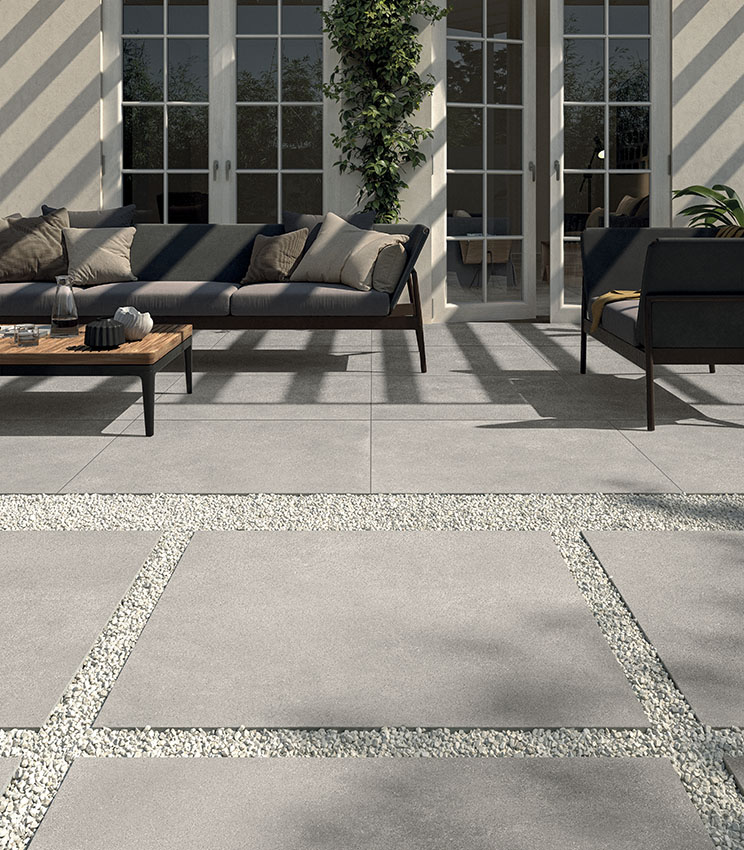 Blick auf einen Patio mit moderner Sitzecke aus Sofa und Sessel. Bodenbelag aus 20 mm starkem Feinsteinzeug in Betonoptik, im Klebeverfahren verlegt. Im Vordergrund ein Outdoor-Weg aus großformatigen Keramikplatten, trocken auf Kies verlegt. | Kollektion Formula von Panaria Ceramica