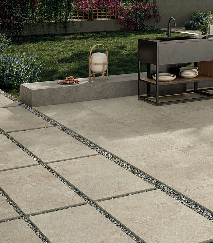 Blick auf einen Poolbereich mit einer modernen Außenküche und einem grünen Garten im Hintergrund. Der Feinsteinzeugboden in Steinoptik in Sandfarbe ist rund um den Pool mit Klebstoff verlegt und im Vordergrund trocken auf Kies als Gehweg verlegt | Revel Kollektion von Panaria Ceramica