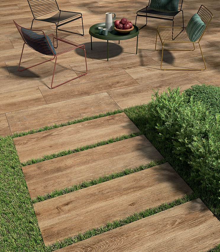 Blick auf einen Patio mit modernen Drahtgestell-Stühlen und einem 20 mm starken Feinsteinzeugboden in Holzoptik, mit Klebstoff verlegt. Im Vordergrund beginnt ein Gehweg aus keramischen Außendielen, trocken auf Rasen verlegt | Kairos Kollektion von Panaria Ceramica