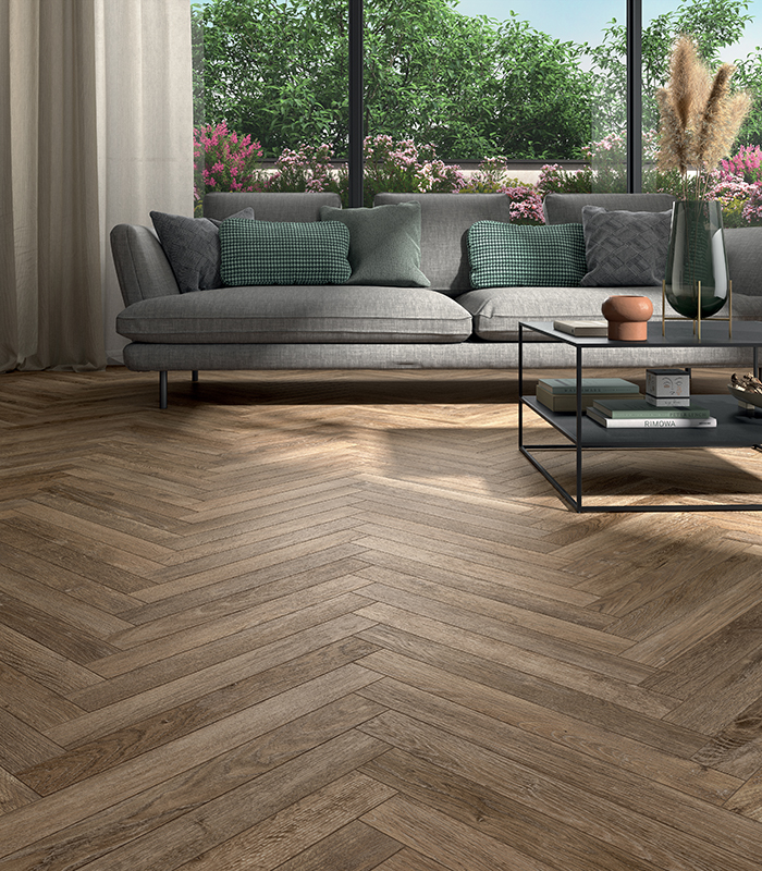 Scorcio di living con ampia porzione di pavimento in gres effetto legno rustico fortemente stonalizzato, moderno divano grigio con tavolino da caffè e, alle spalle, vetrata che dà su un giardino rigoglioso.