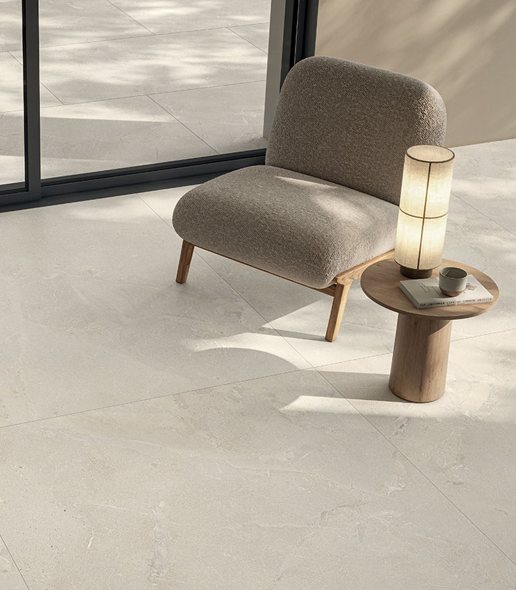 Heller, moderner Wohnbereich mit gepolstertem Sessel, Holztisch und Feinsteinzeugboden in heller Steinoptik, der sich nahtlos in den Außenbereich fortsetzt.