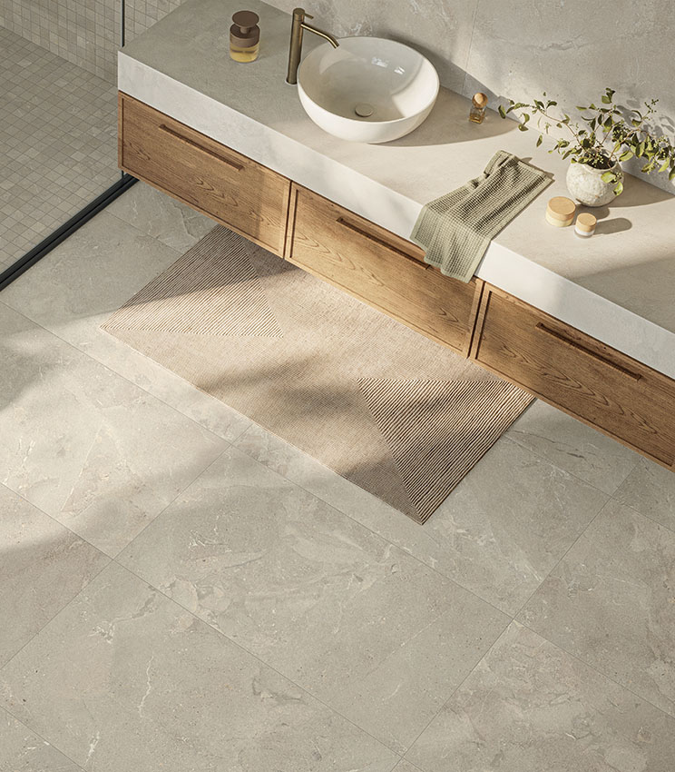 Modernes Badezimmer mit Oberflächen aus den Kollektionen Revel Dune, Mosaico Dune und Workshop Jade, geprägt von hellen Boden- und Wandflächen in Steinoptik. Der Raum verfügt über einen langen Waschtisch aus Holz, eine helle Ablage mit weißem Aufsatzwaschbecken, goldfarbene Armaturen, einen geometrischen Teppich und eine Dusche mit abgestimmtem Mosaik.