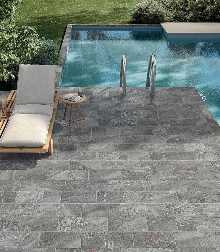 Poolbereich mit Feinsteinzeugboden in grauer Steinoptik, Sonnenliege und Poolleiter für ein elegantes, natürlich wirkendes und widerstandsfähiges Ambiente.